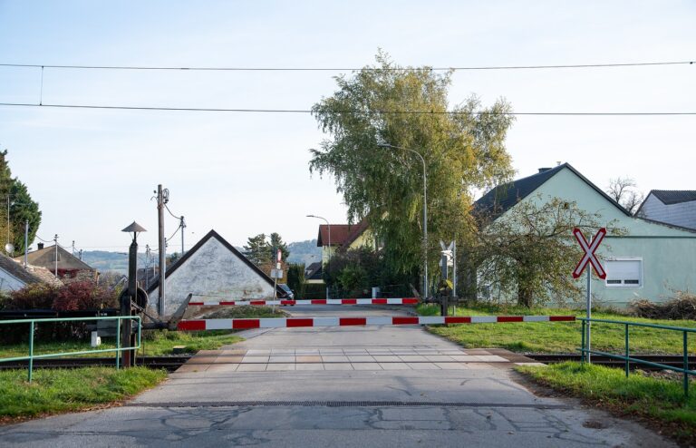 A lowered red-and-white level-crossing barrier across a quiet village road, with houses, a tall tree and a railway crossing sign