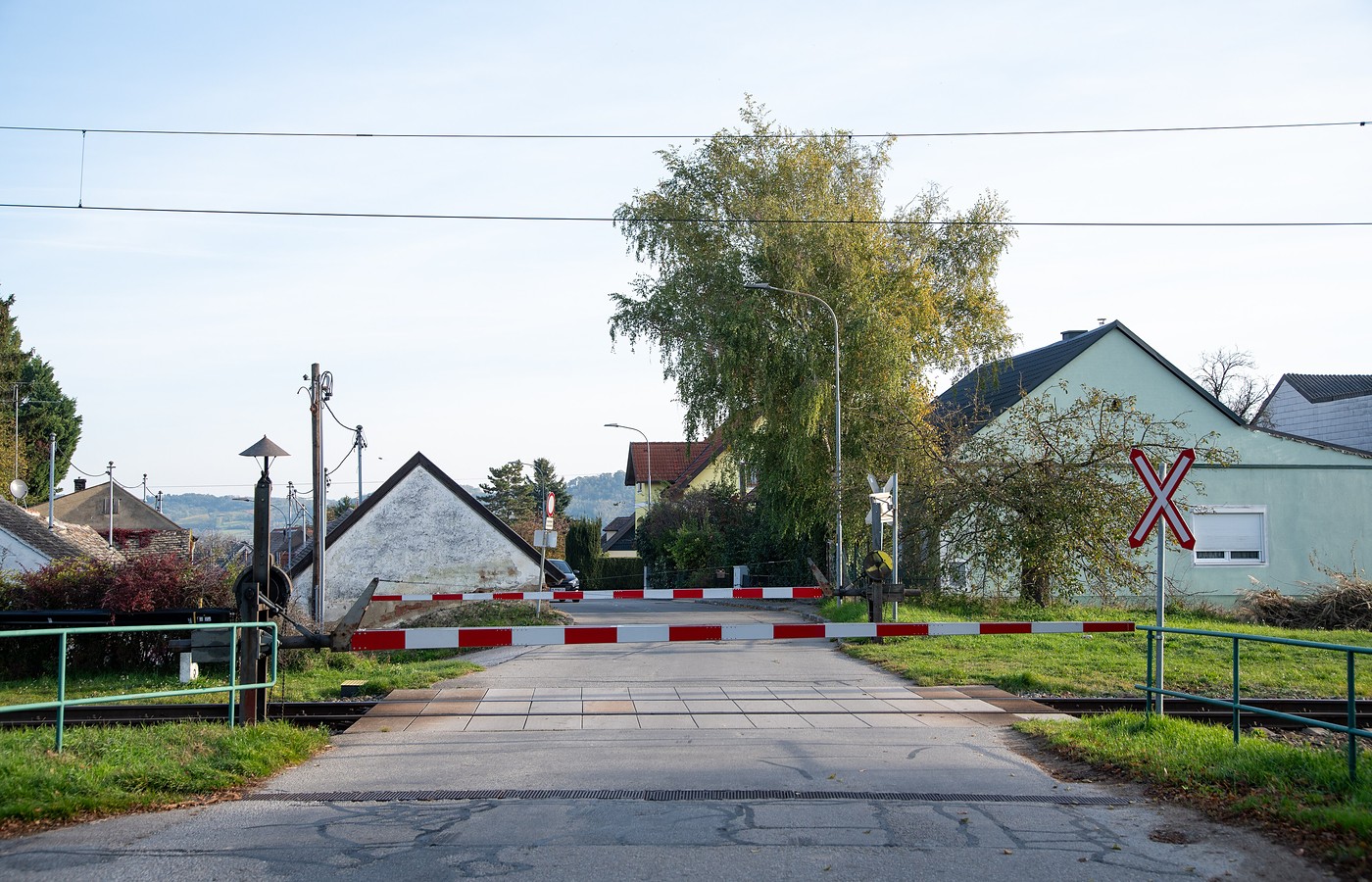 A lowered red-and-white level-crossing barrier across a quiet village road, with houses, a tall tree and a railway crossing sign