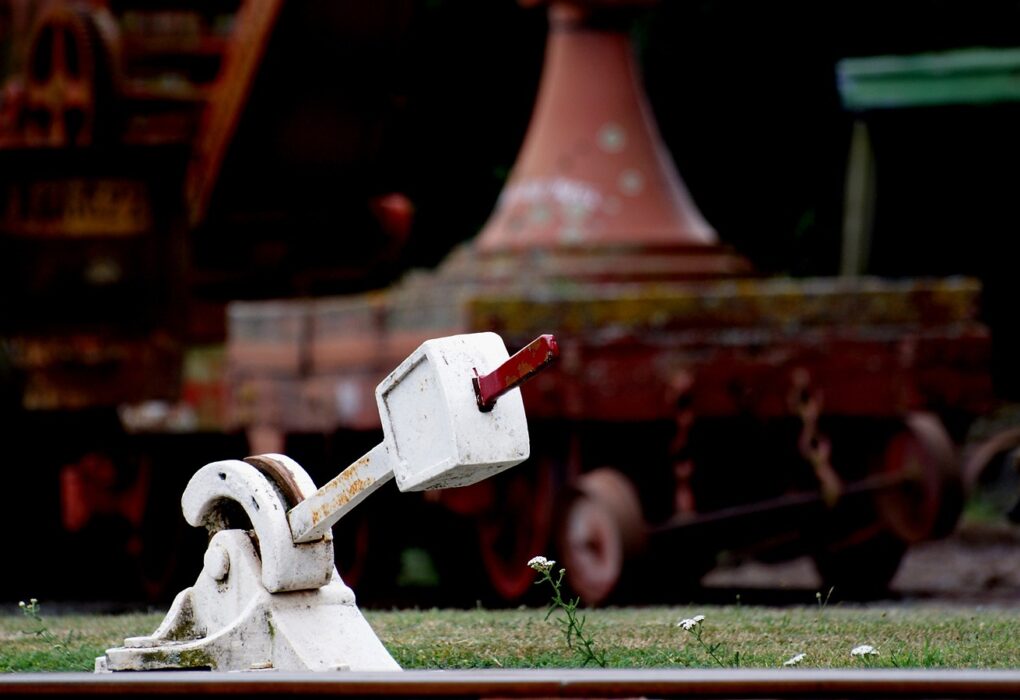A white railway points lever with a red handle beside the rail, old rolling stock blurred behind