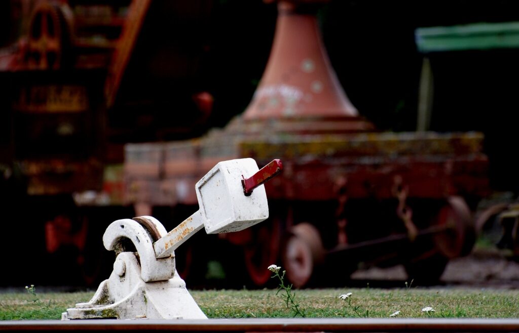 A white railway points lever with a red handle beside the rail, old rolling stock blurred behind