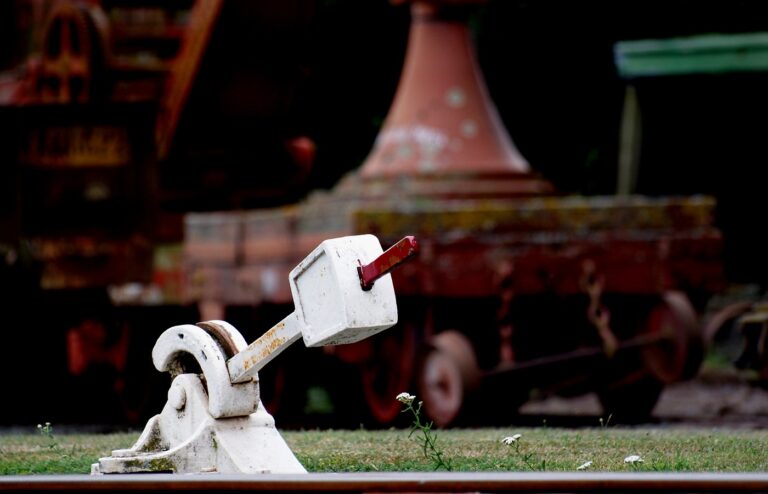 A white railway points lever with a red handle beside the rail, old rolling stock blurred behind