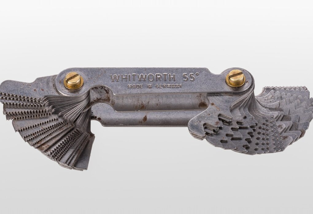 A steel thread pitch gauge, a folded fan of metal blades with different tooth patterns, on a plain light grey background